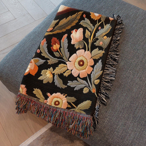 A luxury woven jacquard blanket in three sizes featuring an all-over wild herb botanical pattern of stylized blossoms and foliage in muted gold, olive, lavender, and pewter on a deep black cotton ground; finely twisted multicolor fringe edges complete the artful tapestry weave, ideal as a cozy sofa throw or dramatic wall hanging.